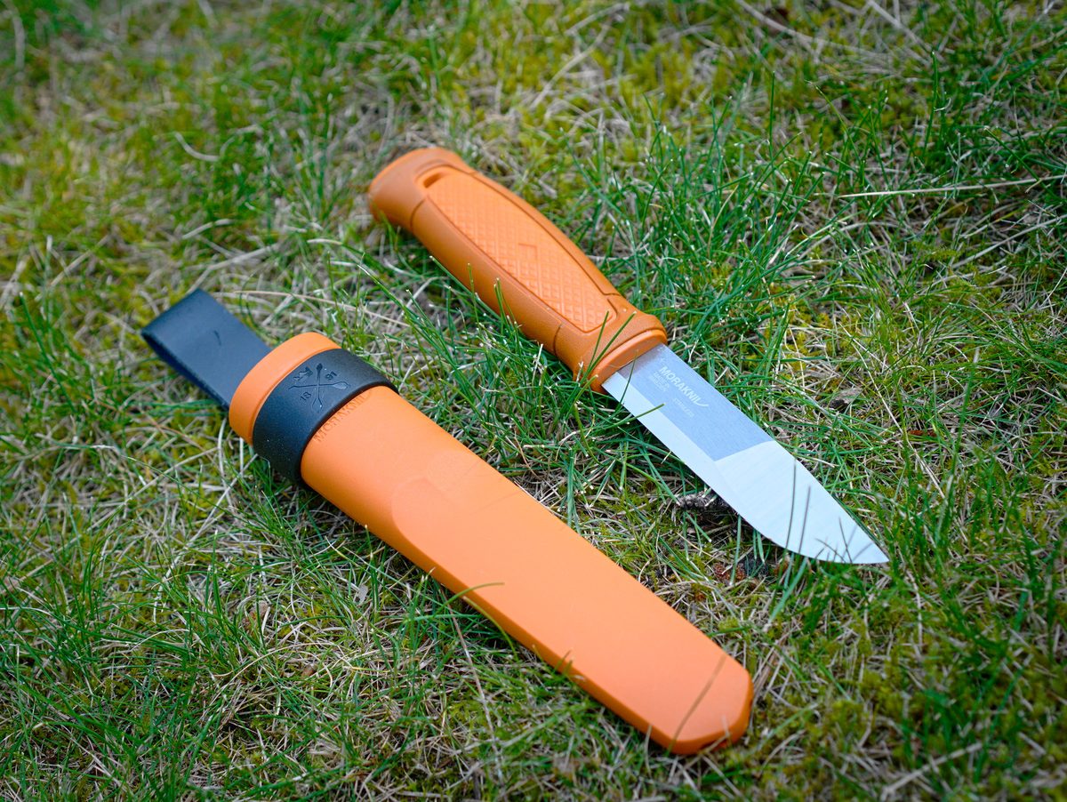 Morakniv Kansbol fixed-blade knife with orange handle beside its orange plastic sheath, on grass