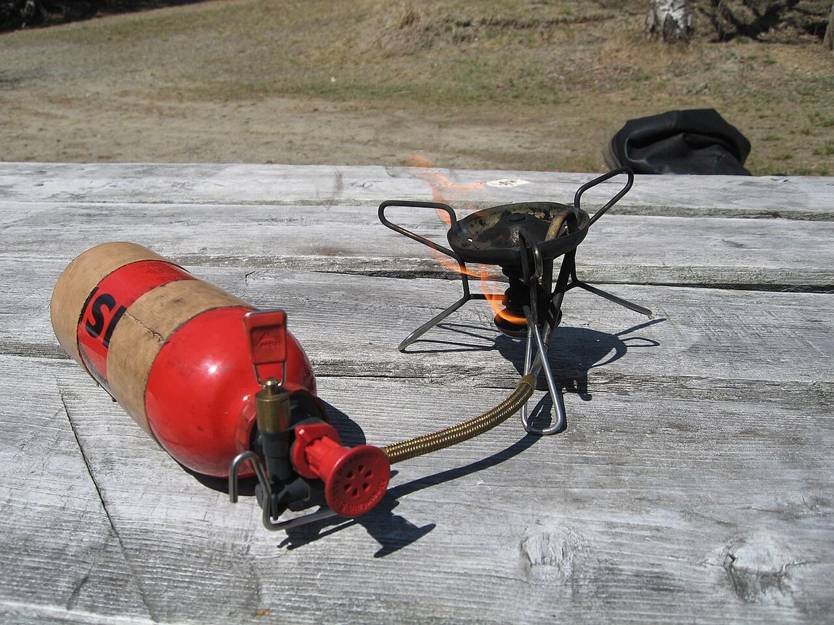 An MSR WhisperLite stove being primed on a picnic table, with a yellow priming flame under the burner and the fuel bottle attached