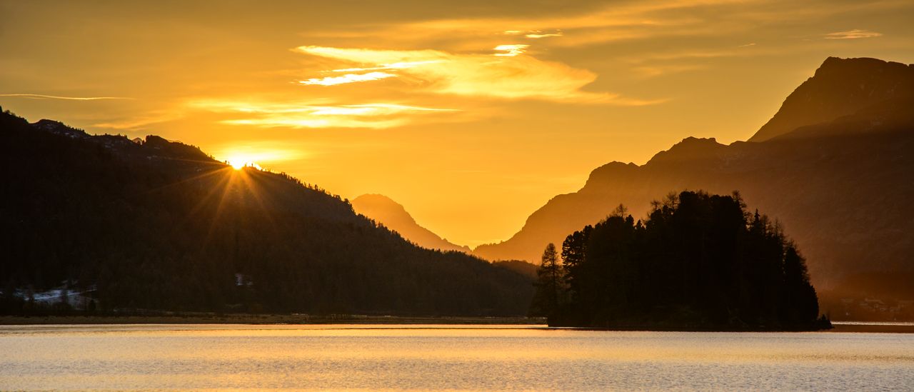 Golden sunset over a quiet alpine lake, the sun dropping behind forested mountain ridgelines